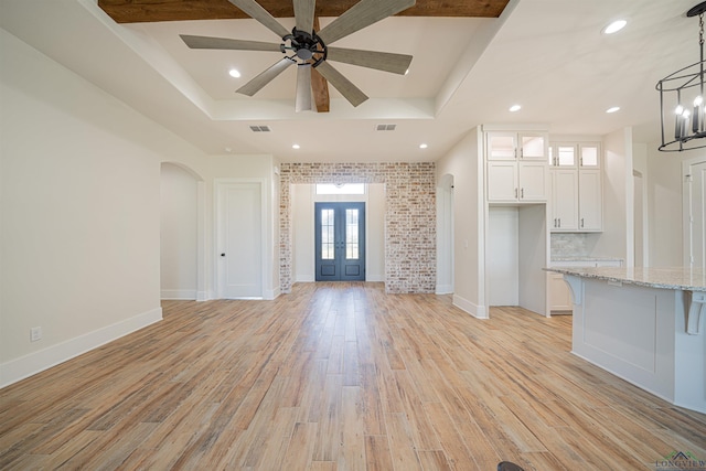 unfurnished living room with french doors, a raised ceiling, light hardwood / wood-style flooring, brick wall, and ceiling fan with notable chandelier