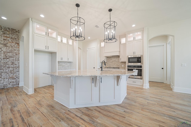 kitchen featuring white cabinetry, built in microwave, stainless steel oven, light stone counters, and a kitchen island with sink