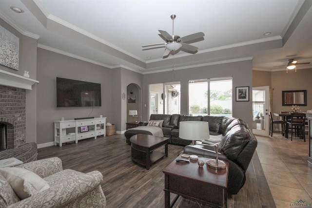 living room featuring dark hardwood / wood-style floors, ornamental molding, ceiling fan, a tray ceiling, and a brick fireplace