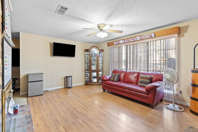 living room with a textured ceiling, light wood-style flooring, visible vents, baseboards, and a ceiling fan