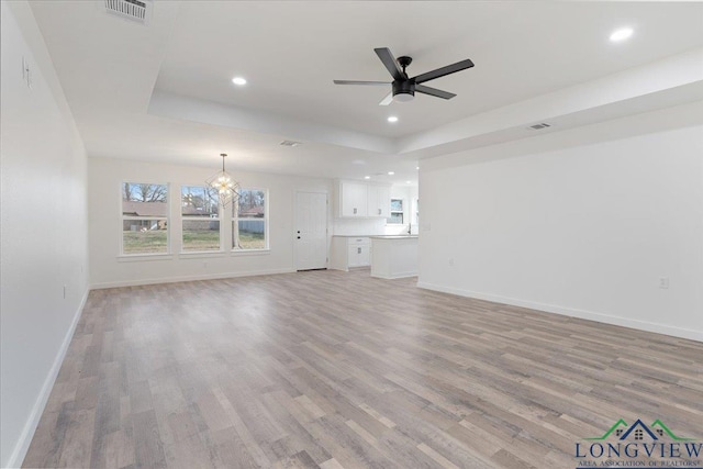 unfurnished living room featuring ceiling fan with notable chandelier, plenty of natural light, light hardwood / wood-style floors, and a raised ceiling