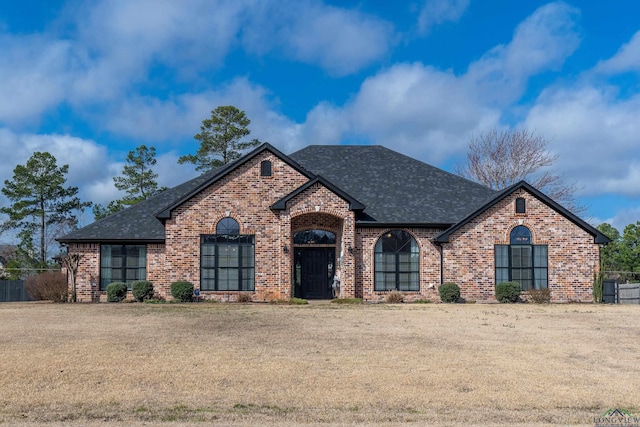 french provincial home featuring roof with shingles, a front yard, and brick siding