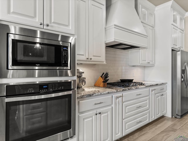 kitchen with premium range hood, white cabinetry, stainless steel appliances, light stone countertops, and light wood-type flooring