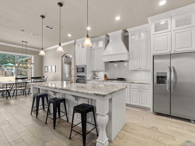 kitchen featuring stainless steel appliances, premium range hood, white cabinets, and an island with sink