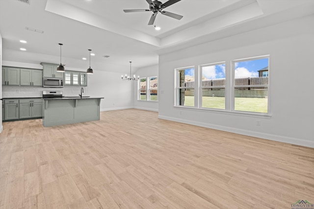 unfurnished living room featuring sink, ceiling fan with notable chandelier, a tray ceiling, and light hardwood / wood-style floors