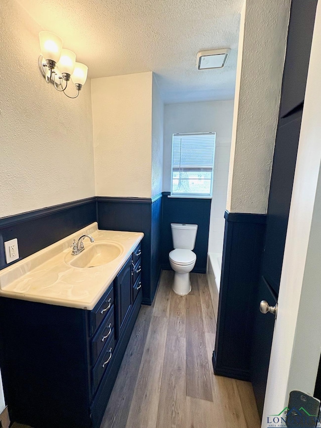 full bath featuring a wainscoted wall, toilet, vanity, wood finished floors, and a textured ceiling
