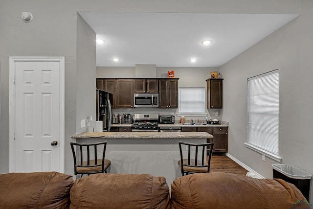 kitchen with light stone countertops, a kitchen bar, dark hardwood / wood-style flooring, dark brown cabinets, and stainless steel appliances