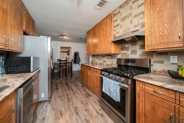 kitchen with appliances with stainless steel finishes, light stone counters, a textured ceiling, light hardwood / wood-style flooring, and tasteful backsplash