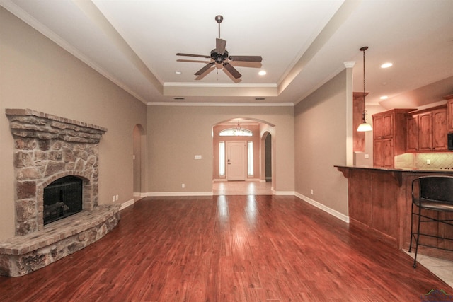 unfurnished living room with a raised ceiling, ceiling fan, dark wood-type flooring, crown molding, and a stone fireplace