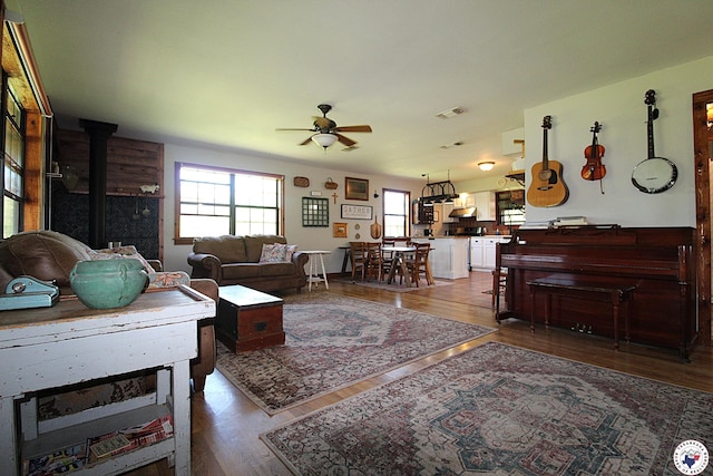 living area featuring plenty of natural light, a ceiling fan, wood finished floors, and a wood stove
