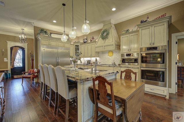 kitchen featuring a center island with sink, appliances with stainless steel finishes, glass insert cabinets, custom exhaust hood, and cream cabinetry