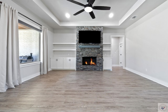 unfurnished living room with a stone fireplace, built in shelves, baseboards, light wood-style floors, and a tray ceiling