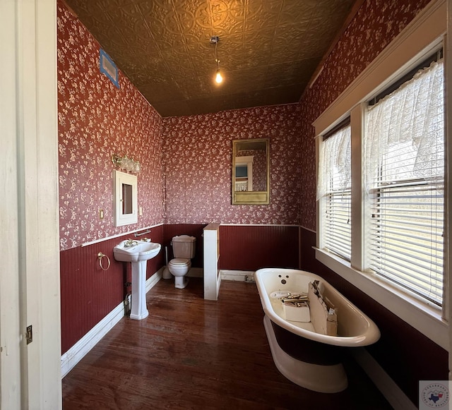 full bath with wallpapered walls, toilet, wood finished floors, a soaking tub, and an ornate ceiling