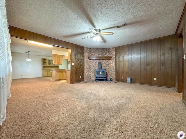 unfurnished living room with a textured ceiling, light colored carpet, ceiling fan, wooden walls, and a wood stove