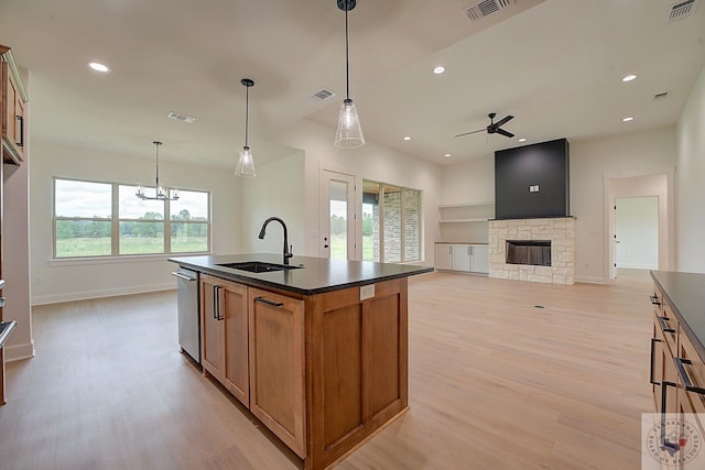 kitchen with a stone fireplace, hanging light fixtures, a kitchen island with sink, stainless steel dishwasher, and sink