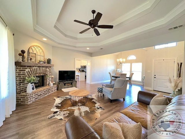 living room featuring ceiling fan with notable chandelier, hardwood / wood-style floors, ornamental molding, a raised ceiling, and a brick fireplace