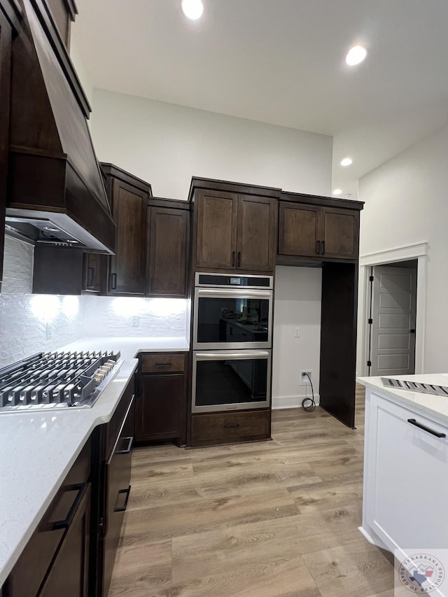 kitchen with custom exhaust hood, appliances with stainless steel finishes, decorative backsplash, light wood-type flooring, and dark brown cabinetry