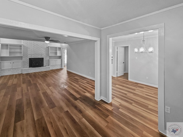 unfurnished living room featuring crown molding, built in features, dark wood-type flooring, a fireplace, and ceiling fan with notable chandelier