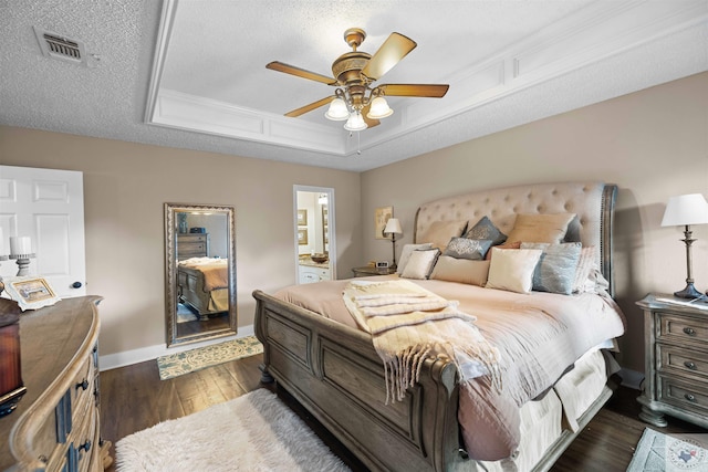 bedroom with a textured ceiling, ensuite bath, dark wood-type flooring, ceiling fan, and a tray ceiling