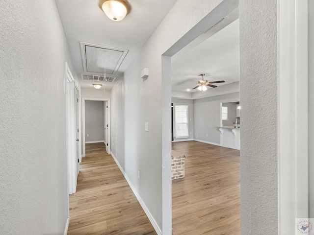 corridor featuring attic access, visible vents, baseboards, a textured wall, and light wood-type flooring