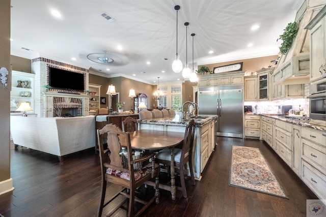 kitchen featuring appliances with stainless steel finishes, cream cabinetry, decorative light fixtures, light stone counters, and a center island with sink