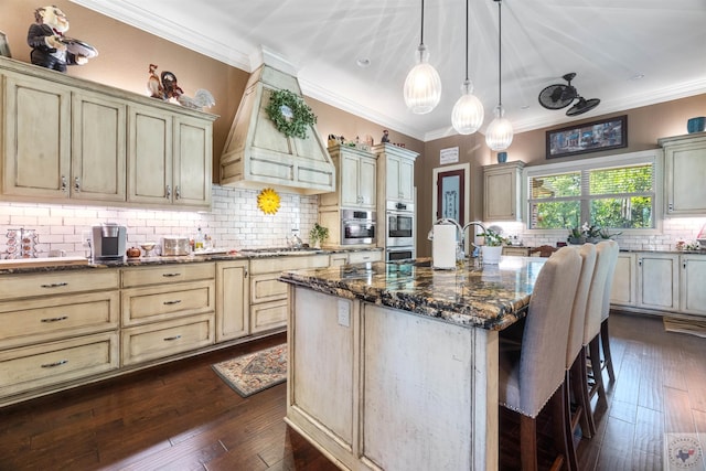 kitchen featuring custom exhaust hood, dark stone counters, an island with sink, cream cabinets, and a breakfast bar area