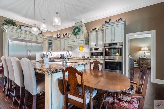 kitchen with pendant lighting, cream cabinets, dark stone counters, a center island with sink, and stainless steel double oven