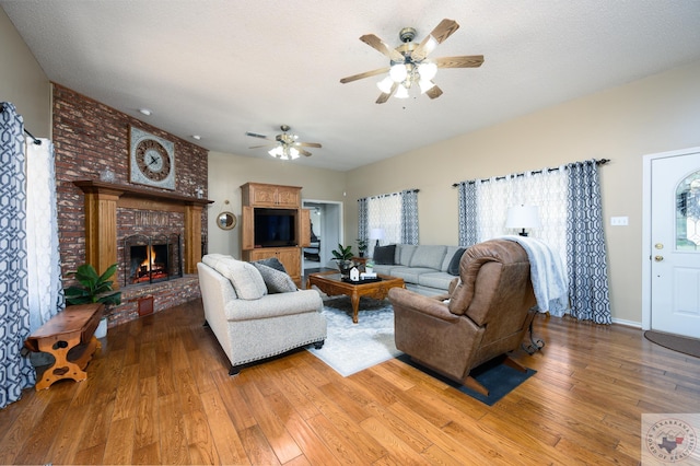 living room with ceiling fan, a textured ceiling, a brick fireplace, and light wood-style flooring