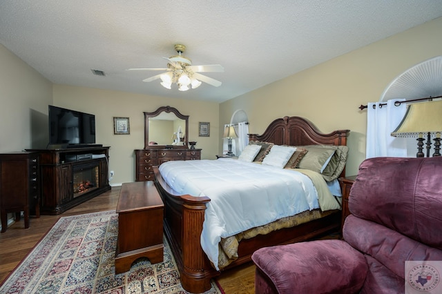 bedroom featuring visible vents, a textured ceiling, a glass covered fireplace, wood finished floors, and ceiling fan