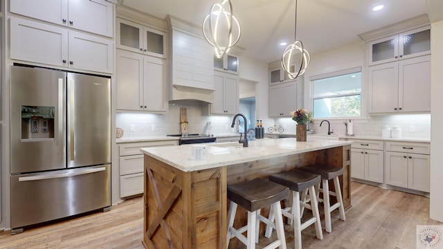 kitchen featuring stainless steel refrigerator with ice dispenser, light hardwood / wood-style flooring, hanging light fixtures, light stone countertops, and an island with sink