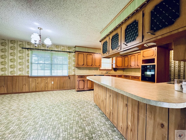 kitchen with a notable chandelier, oven, kitchen peninsula, pendant lighting, and wood walls