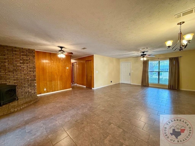 unfurnished living room with a textured ceiling, wood walls, ceiling fan with notable chandelier, and a wood stove