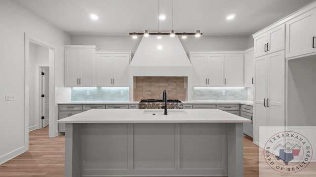 kitchen featuring stainless steel gas cooktop, light countertops, light wood-style floors, an island with sink, and premium range hood