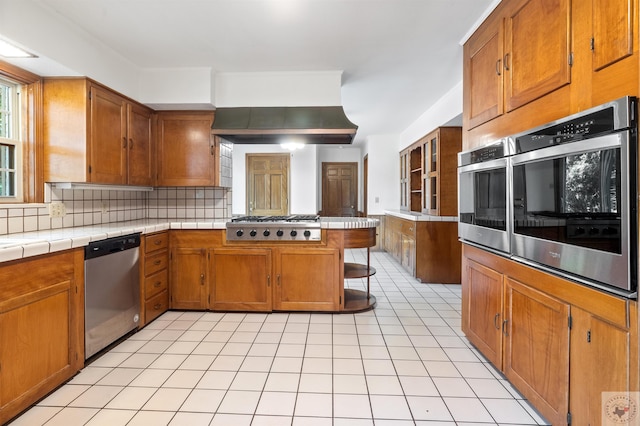 kitchen featuring tile counters, appliances with stainless steel finishes, island range hood, kitchen peninsula, and backsplash