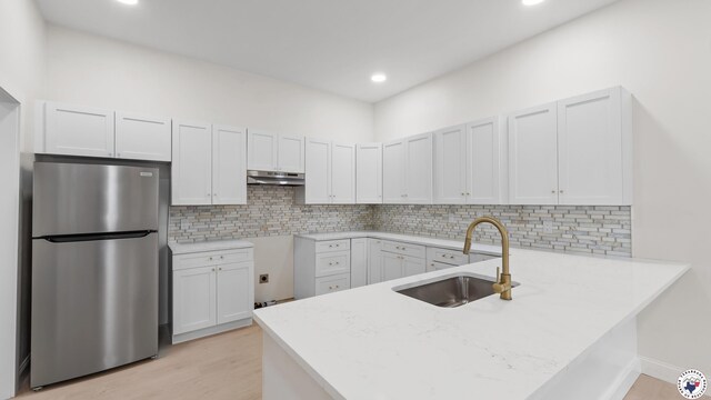 kitchen featuring under cabinet range hood, a sink, freestanding refrigerator, light wood-style floors, and a peninsula