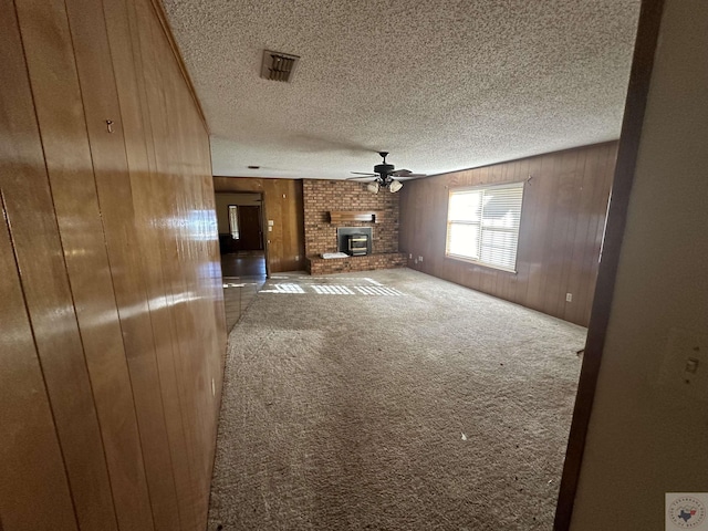 unfurnished living room featuring a textured ceiling, a brick fireplace, wooden walls, carpet, and ceiling fan