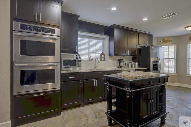 kitchen with dark brown cabinetry, stainless steel appliances, sink, backsplash, and light stone counters