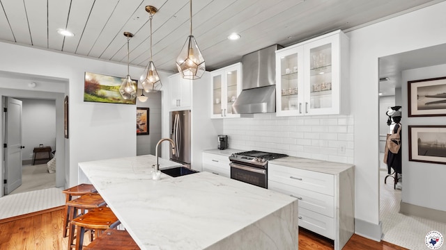 kitchen with light stone counters, a sink, stainless steel appliances, a center island with sink, and wall chimney exhaust hood