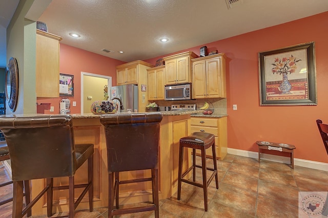 kitchen with kitchen peninsula, light brown cabinetry, appliances with stainless steel finishes, a breakfast bar, and decorative backsplash