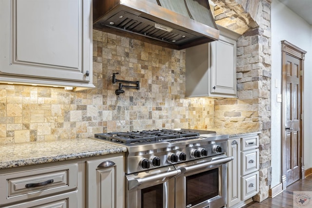 kitchen with light stone counters, dark wood finished floors, range with two ovens, wall chimney exhaust hood, and backsplash