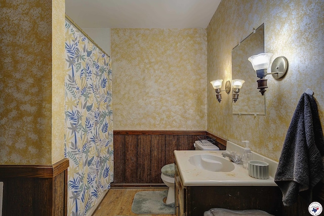 bathroom with wood finished floors, wallpapered walls, wainscoting, vanity, and toilet