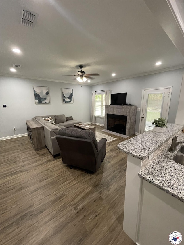 living area featuring dark wood-style flooring, a tiled fireplace, visible vents, a ceiling fan, and ornamental molding