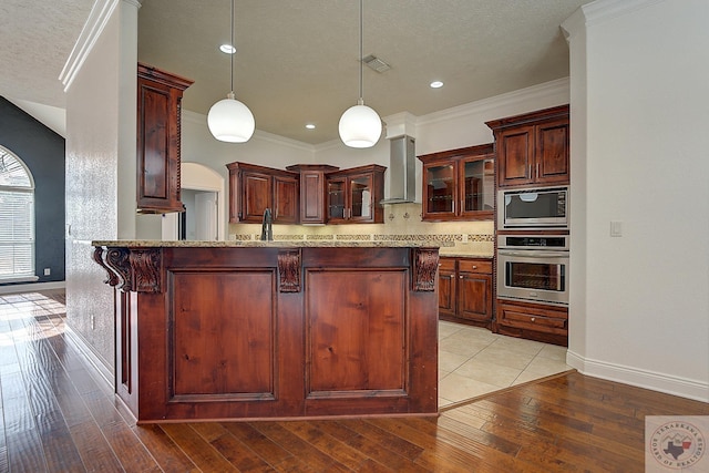 kitchen with kitchen peninsula, wall chimney exhaust hood, decorative light fixtures, ornamental molding, and stainless steel appliances