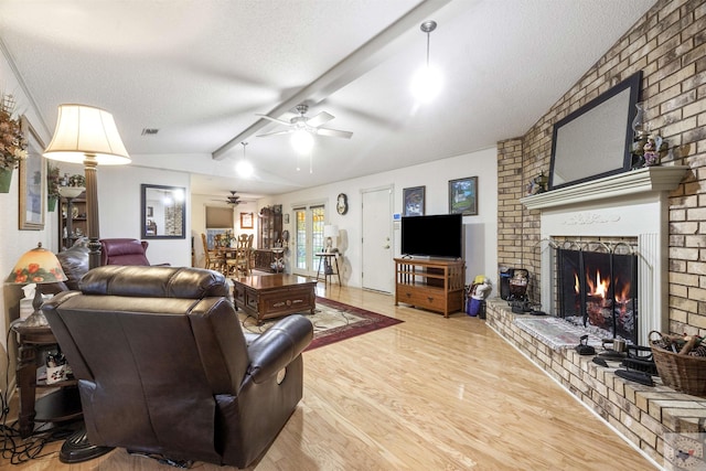 living room featuring ceiling fan, lofted ceiling with beams, a brick fireplace, a textured ceiling, and light hardwood / wood-style floors