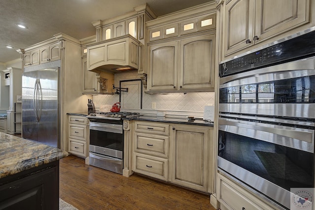 kitchen with appliances with stainless steel finishes, dark stone countertops, ornamental molding, backsplash, and dark hardwood / wood-style floors