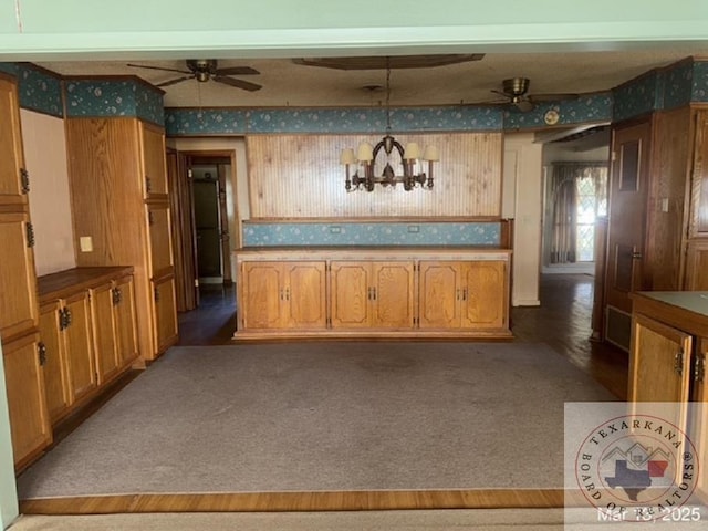 kitchen featuring ceiling fan with notable chandelier, decorative light fixtures, wallpapered walls, brown cabinetry, and carpet flooring