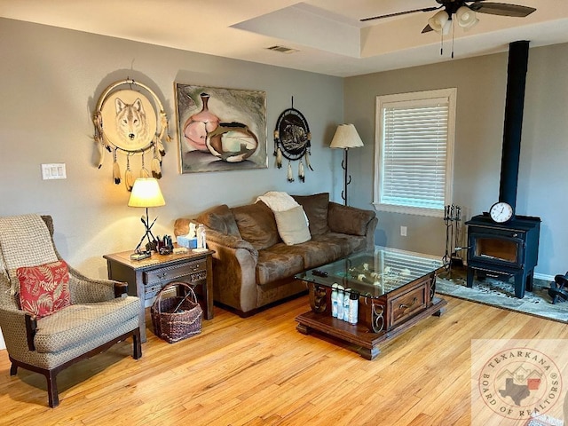 living room featuring ceiling fan, a raised ceiling, light hardwood / wood-style floors, and a wood stove