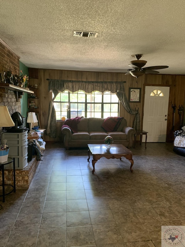 living room with ceiling fan, a brick fireplace, wooden walls, and a textured ceiling