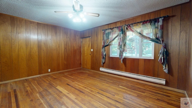 empty room featuring ceiling fan, a baseboard heating unit, hardwood / wood-style floors, a textured ceiling, and wooden walls