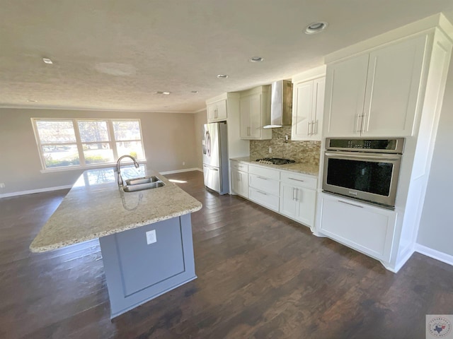 kitchen with appliances with stainless steel finishes, white cabinetry, wall chimney range hood, sink, and an island with sink